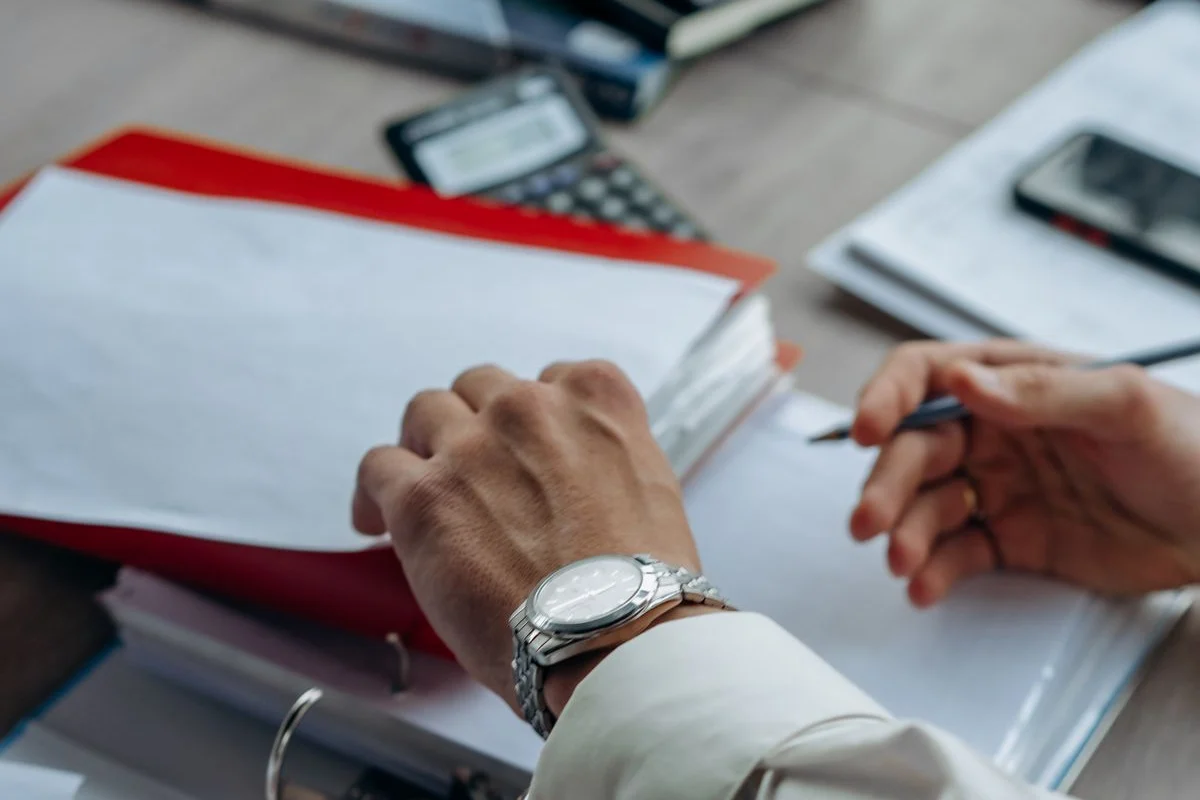 赤いファイルと書類に手を置く手元。「PMO支援についてよくある質問」の話題のイメージ
