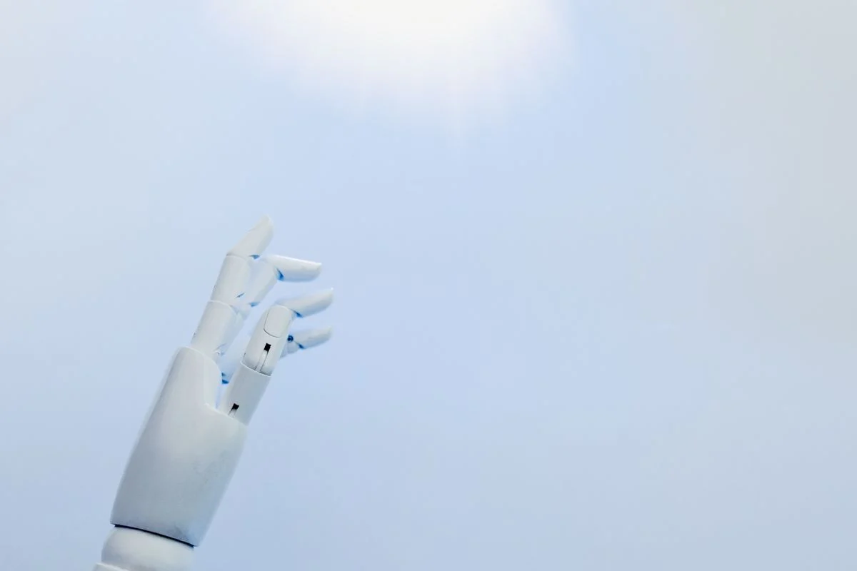 淡い青を背に上へ伸ばす白いロボットの手。AIの出力を確認する層を考える話題のイメージ