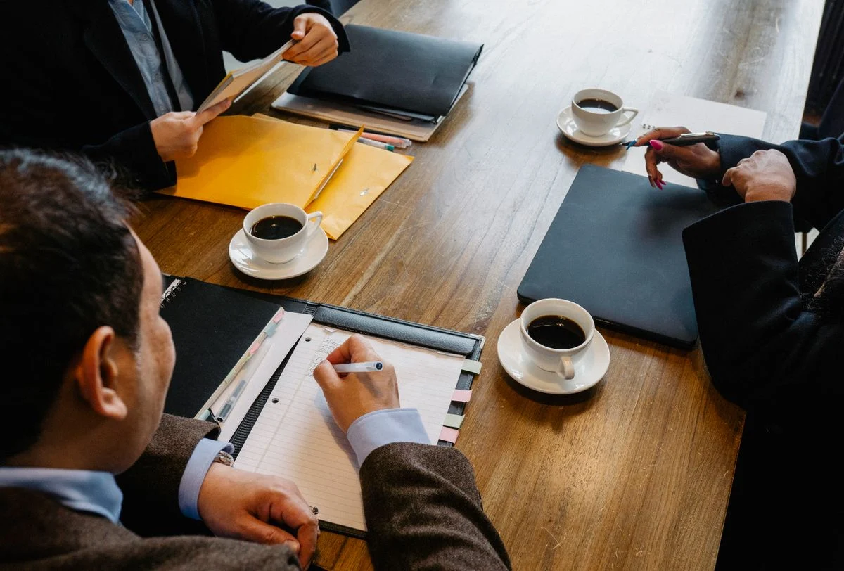 書類とコーヒーカップを前に打ち合わせる人々。「PMO会社に関するよくある質問」の話題のイメージ
